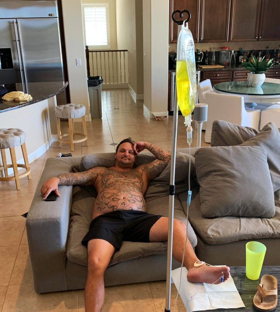 A man with tattoos relaxing on a large grey sofa during a mobile IV infusion session in a modern home.