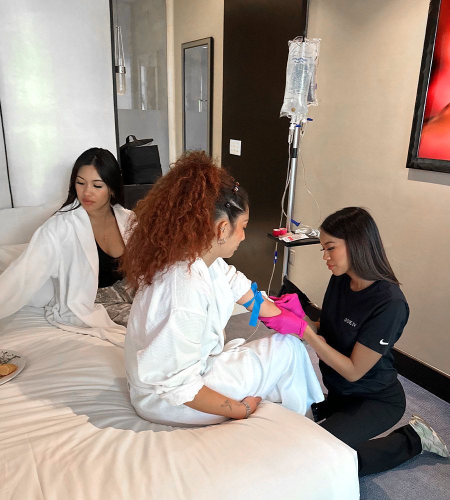 Professional mobile IV therapy nurse in a black uniform kneeling to administer a vitamin infusion to a client wearing a white robe, showcasing high-quality in-room hotel service and care.