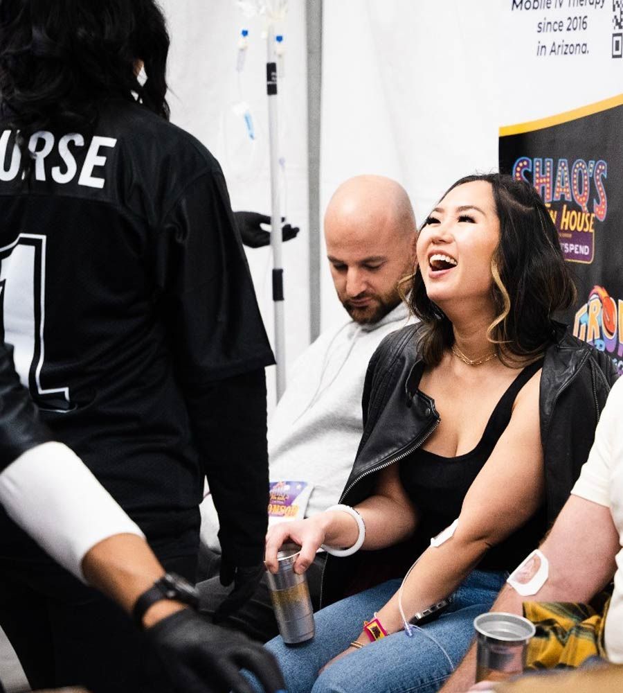 A smiling person holds a drink while sitting near a man in a white shirt, with a staff member in a black jersey nearby.