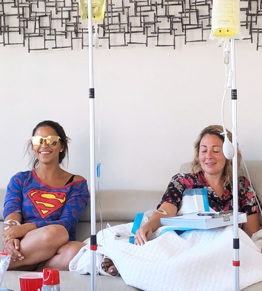 Two people receiving IV drips, smiling, on a sofa. One wears sunglasses and Superman shirt. The other is using headphones.