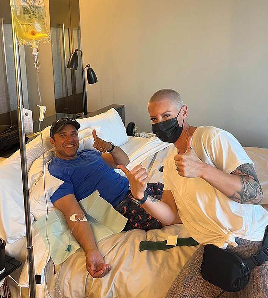 A man in a hotel bed receives a mobile IV therapy drip infusion for travel recovery while giving a thumbs-up next to the masked female provider.
