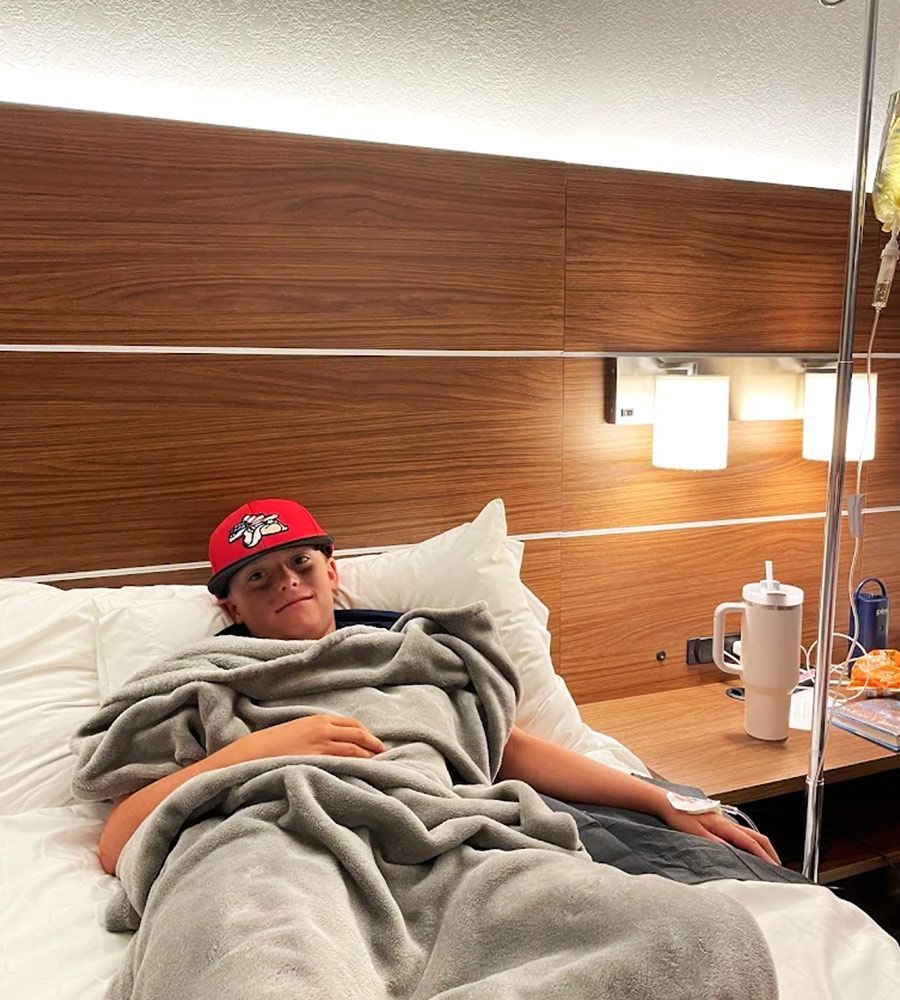 Person in a hospital bed with an IV drip, wearing a baseball cap and wrapped in a blanket.