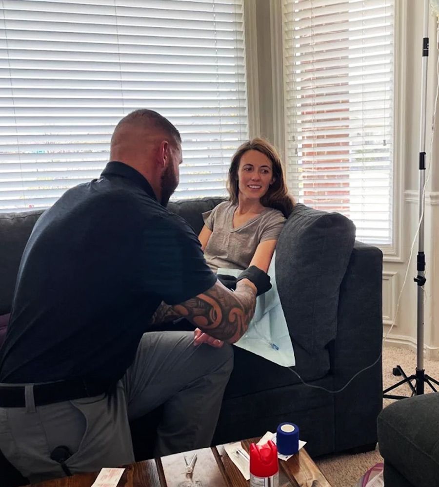 A woman lounging on a bed in a hotel room during a mobile IV hydration session with medical vials in the foreground.