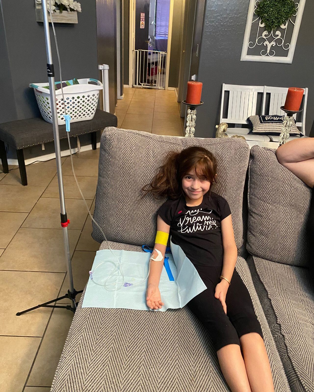 A child sits on a gray sofa receiving an IV infusion, with the fluid bag hanging from a medical stand nearby.