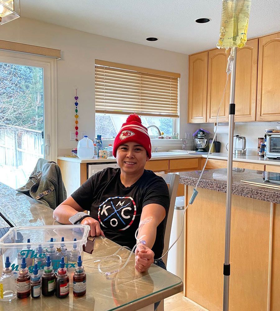 A woman wearing a beanie receiving a wellness IV vitamin treatment at her kitchen table.