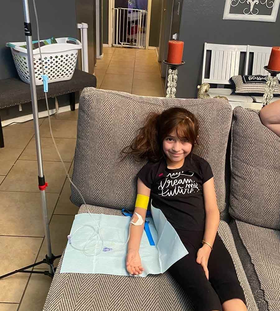 Smiling young girl with long hair relaxing on a couch while receiving mobile IV therapy at home.