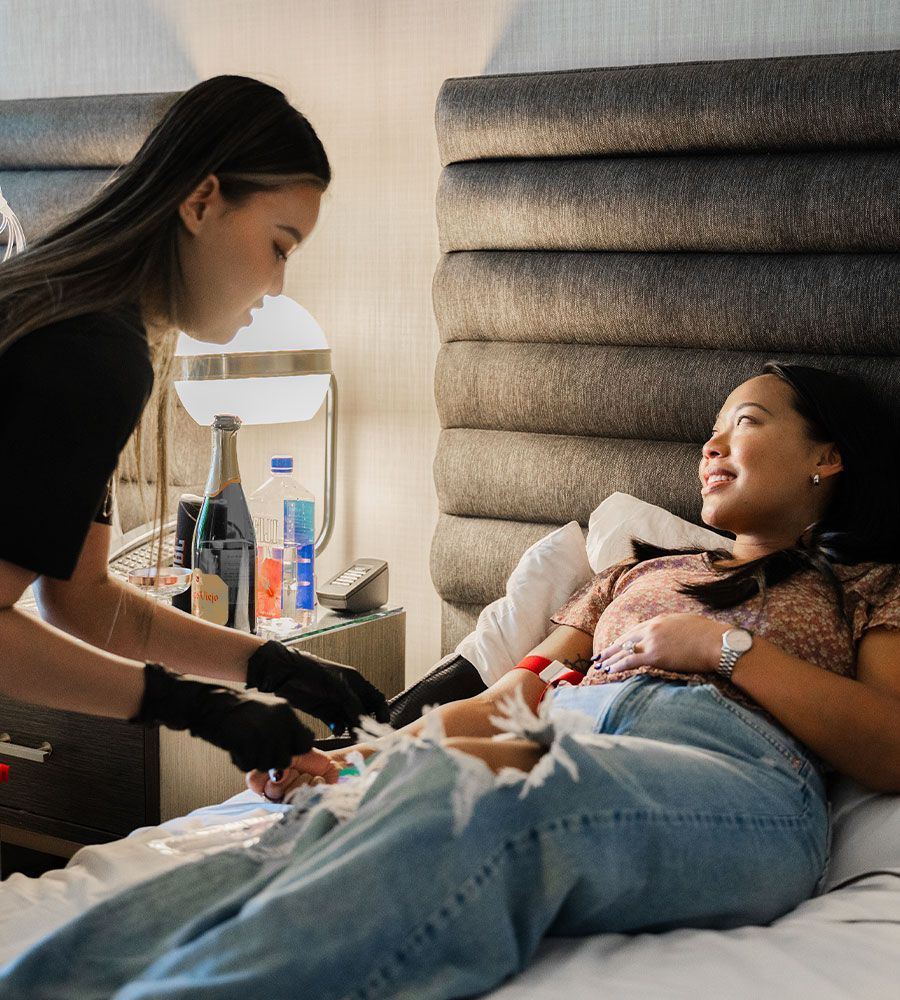Client smiling and relaxing in a hotel bed while receiving mobile IV hydration therapy from a nurse, with champagne and a laptop nearby, highlighting luxury travel recovery.