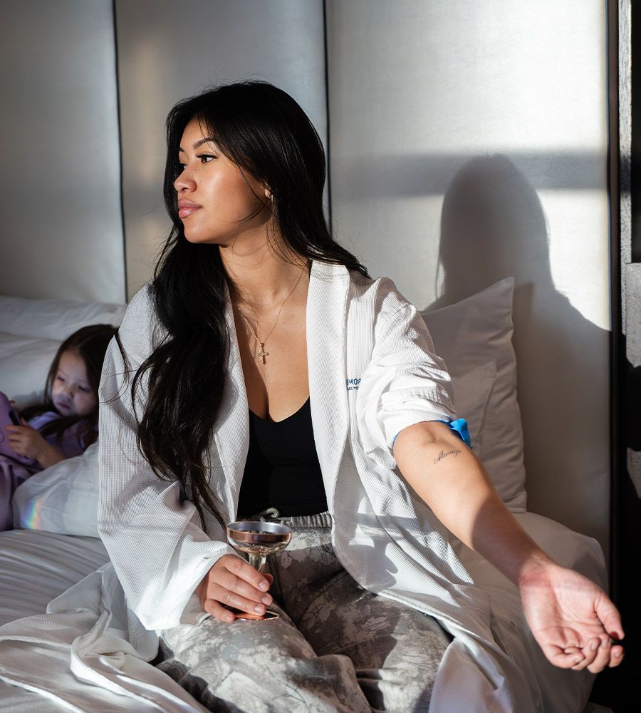 Stylish woman in a white robe relaxing on a hotel bed, holding a glass of champagne and receiving mobile IV therapy, with her daughter playing in the background, showcasing luxury wellness on demand.