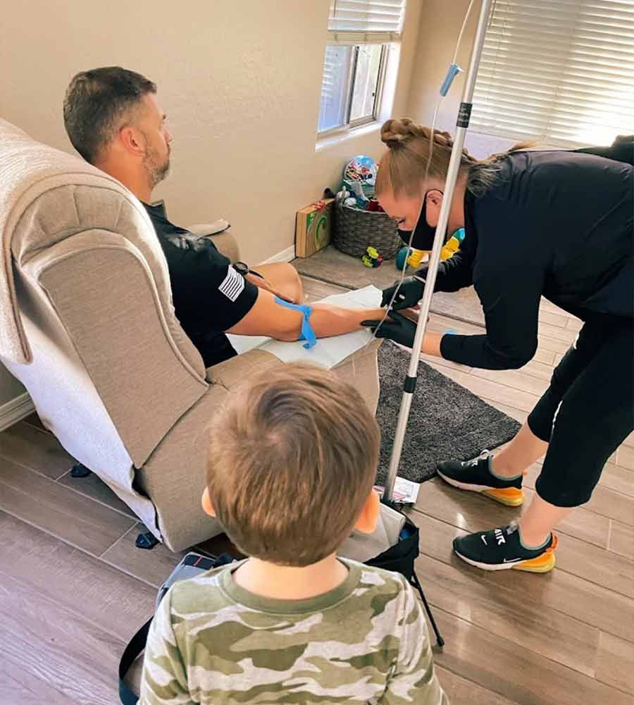 Nurse administering an IV treatment to a male patient sitting in a recliner at home, with a small child watching nearby.