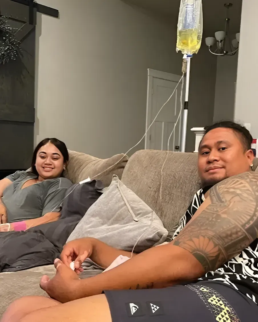 A person with tattoos on their arm sits on a couch receiving IV fluids, with another person sitting nearby smiling.