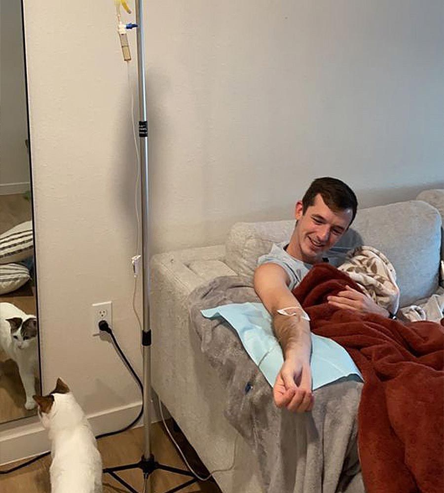 Young man smiling while receiving a mobile IV infusion treatment at home, comfortably tucked in with a blanket on his couch while his cat watches from the floor.