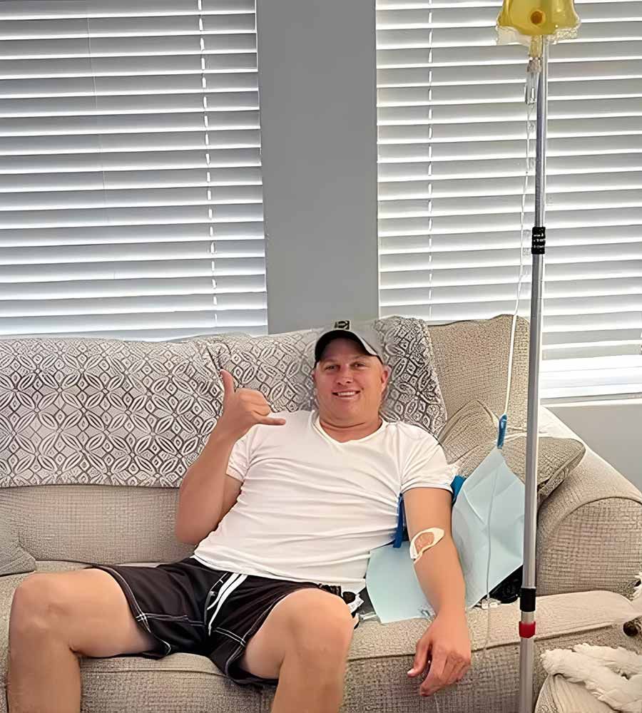 A happy man sits on his couch at home receiving mobile IV therapy and making a shaka sign, demonstrating comfort and satisfaction with the service.