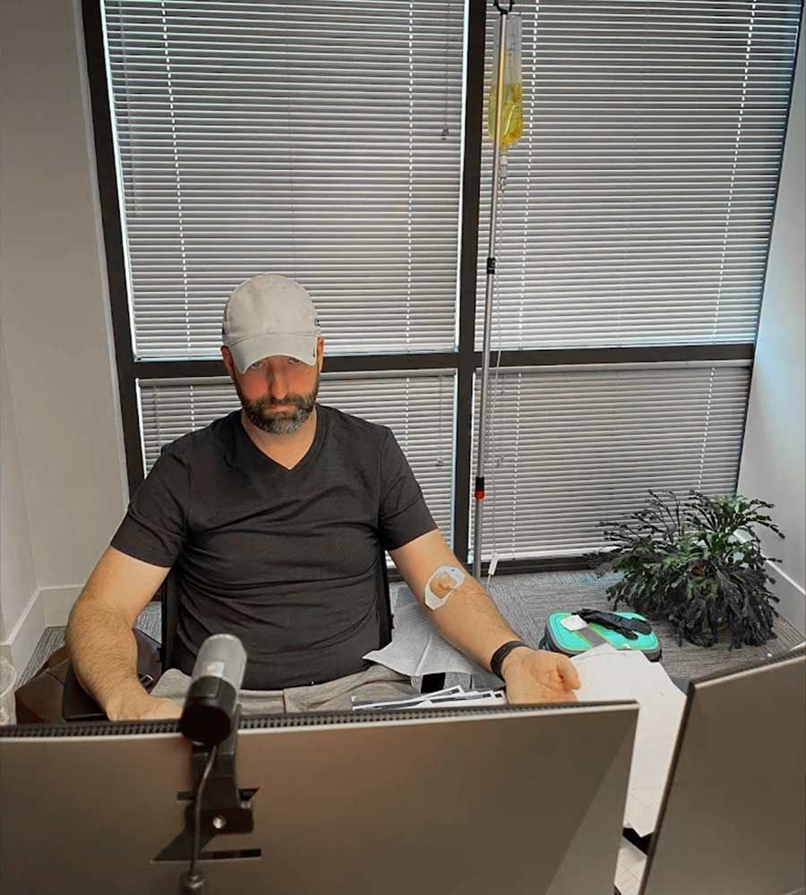 Busy man sitting in a modern office with horizontal blinds receiving a wellness treatment IV drip.