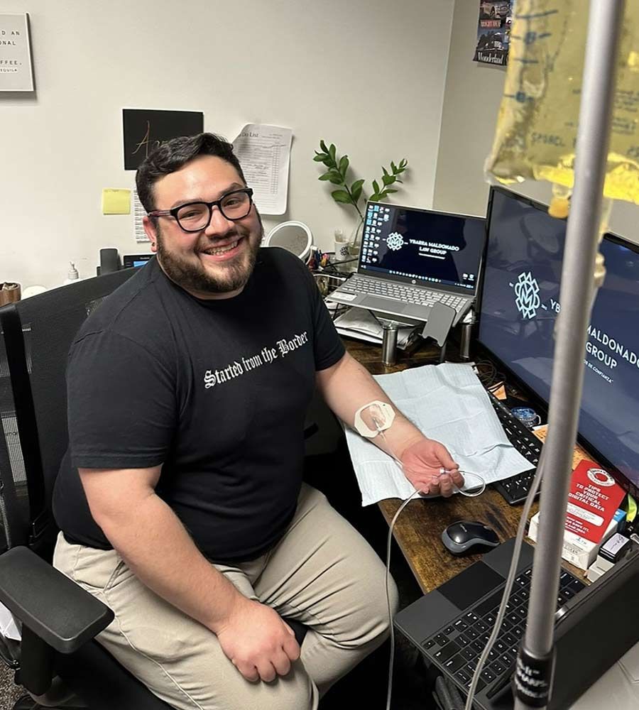 Smiling man with glasses receiving an IV infusion while sitting at his desk with dual monitors in an office.