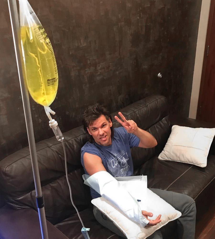Man giving a peace sign while comfortably relaxing on a brown leather couch and receiving mobile IV hydration therapy for quick recovery.