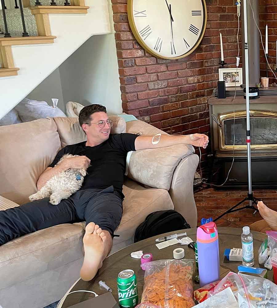 Man on sofa receiving IV, holding a dog, with medical supplies on table.