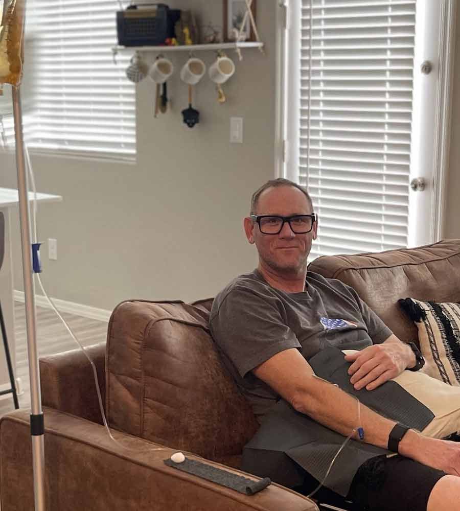 Middle-aged man smiling and relaxing on a brown couch while receiving an at-home IV vitamin infusion.