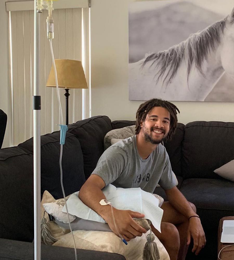 Man on couch receiving IV fluids, smiling at camera. Home setting, IV pole visible.