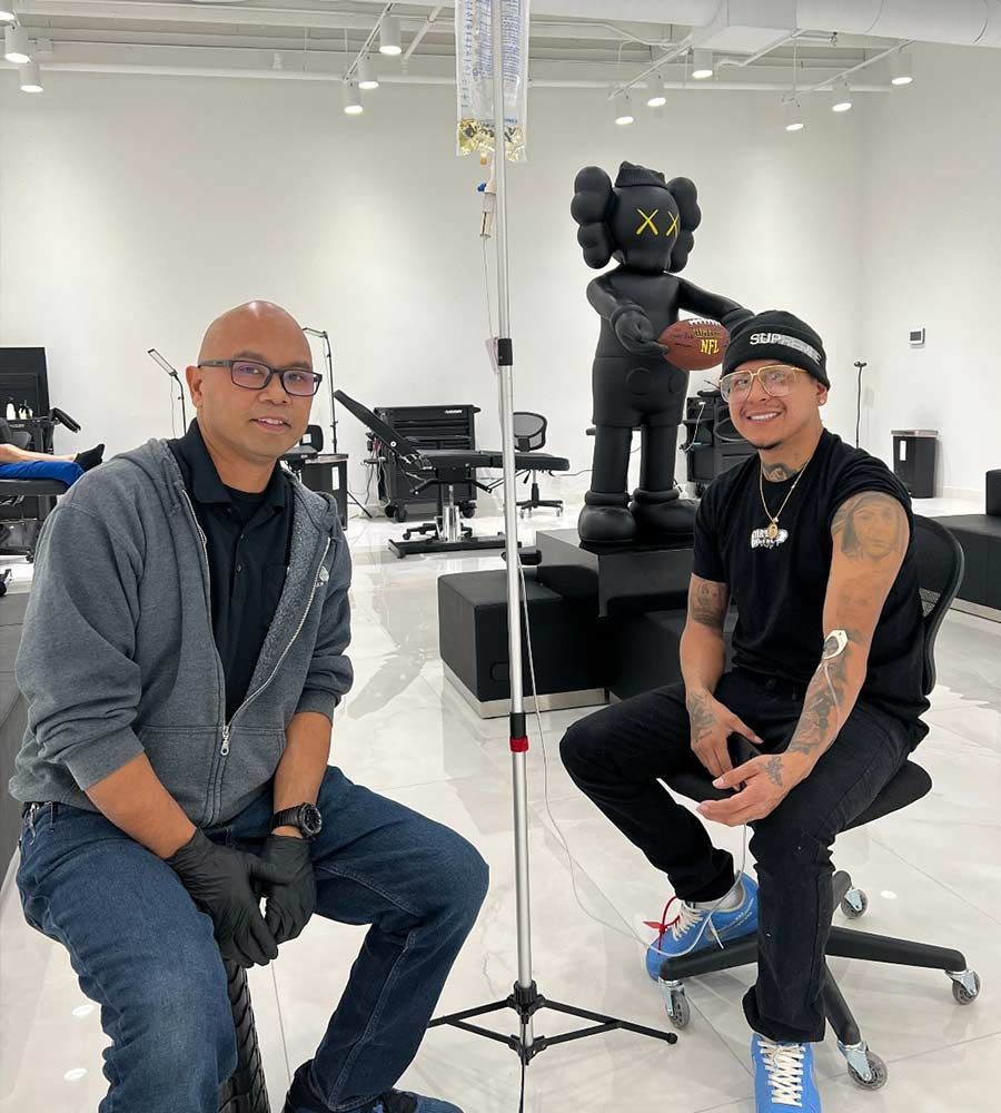 Two people seated near IV drip stand, with modern art in background. Man on right has tattoo.