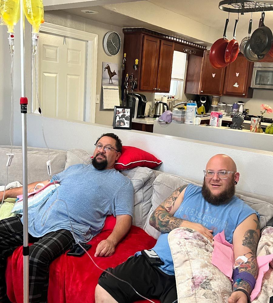 Two men sitting on a grey couch receiving IV drip therapy together in a home setting.