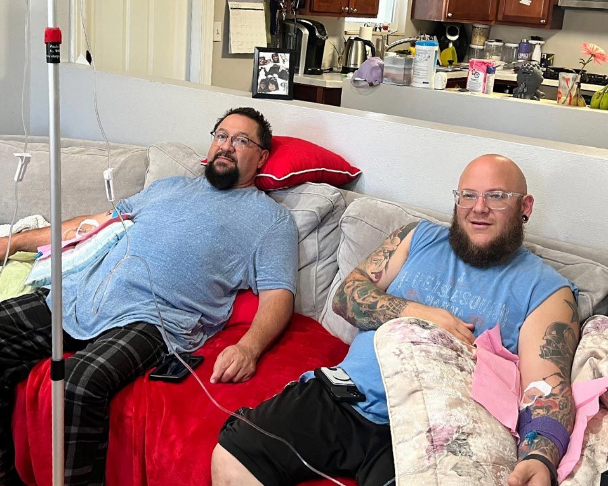 Two people receiving IV fluids on a couch, with IV poles. One has a beard and glasses.