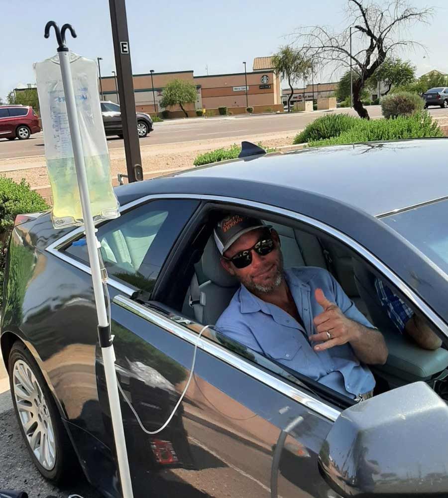 Man in car receiving IV drip, thumbs up. Drip stand outside car. Daytime, sunny.