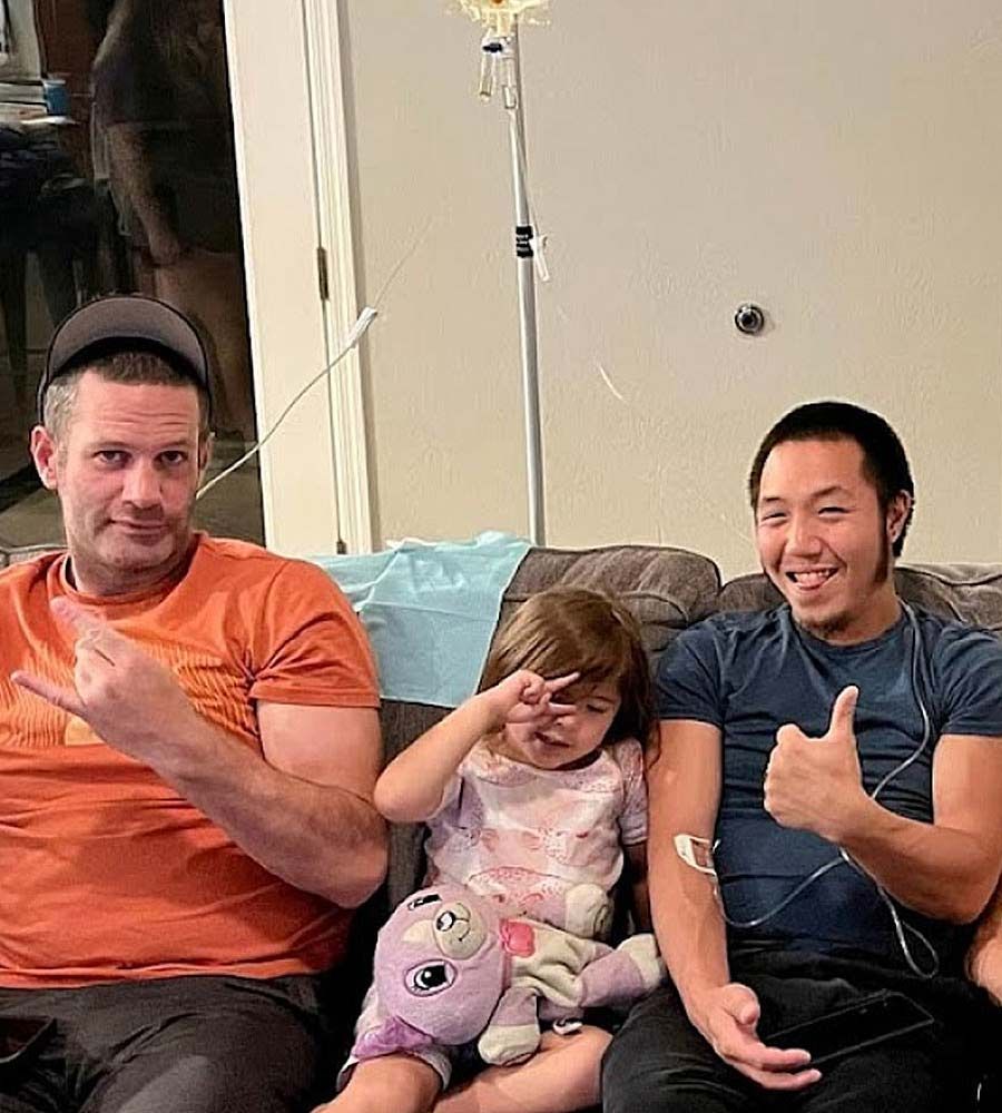 Two men receiving mobile IV hydration therapy on a couch in a dark living room, smiling and giving a thumbs up sign while a child sits between them.