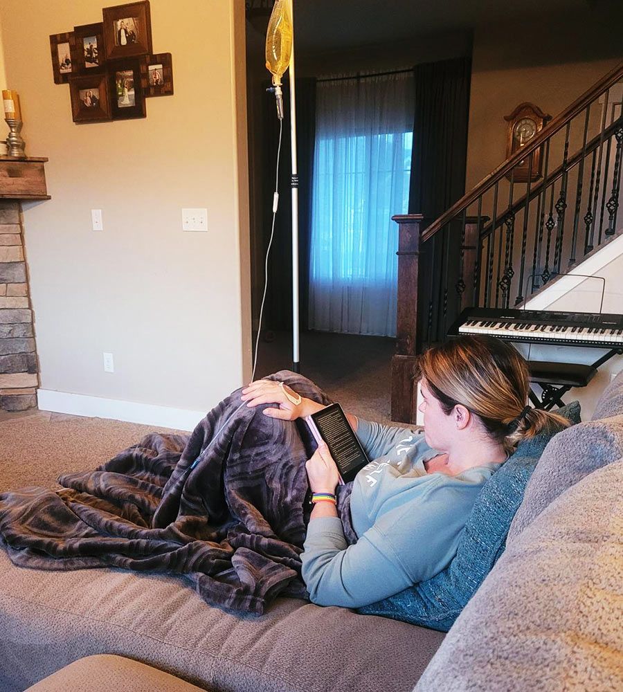A woman resting under a blanket and reading a book on a tablet during an at-home IV therapy session.