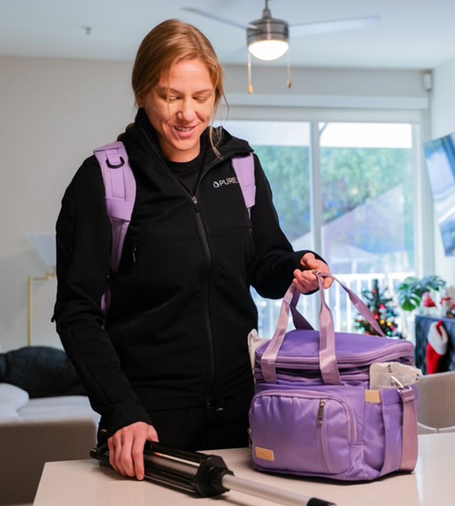 Woman in black jacket with a purple backpack and cooler, holding a travel umbrella indoors.