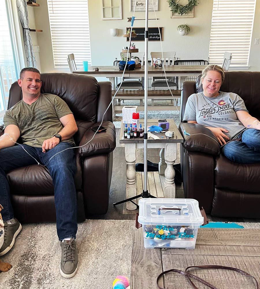 A couple reclining in brown leather chairs receiving at-home mobile IV hydration therapy.