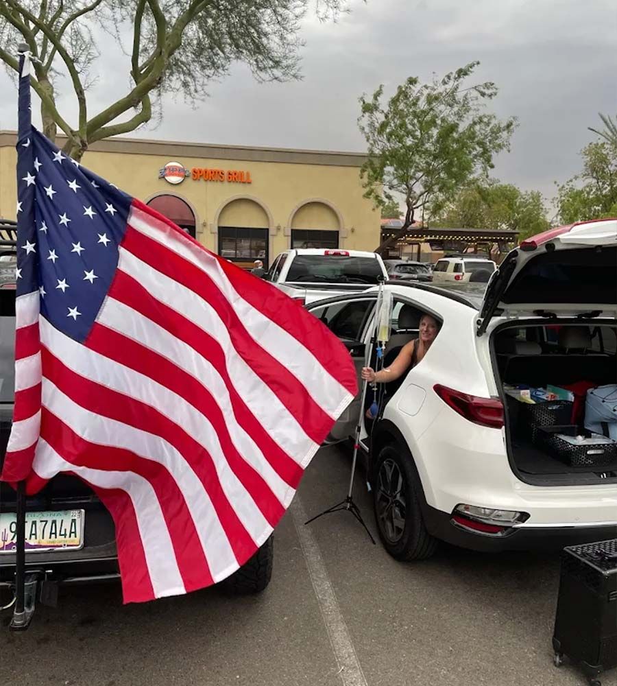 Smiling technician setting up mobile IV therapy equipment next to a white SUV in a parking lot, with a large American flag visible.