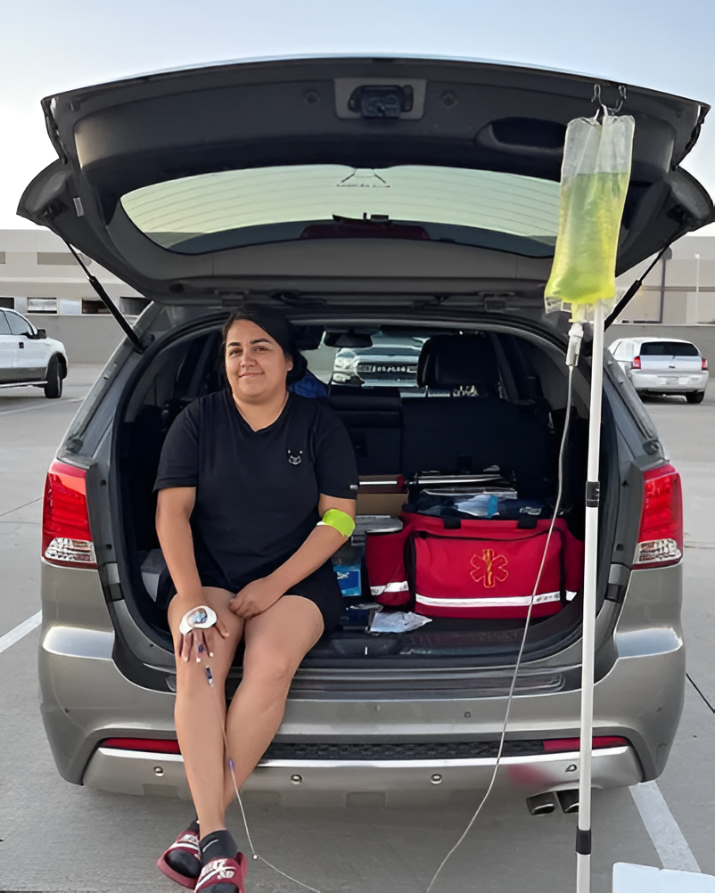 A person sitting in an open car trunk with an IV bag on a stand, wearing an IV line and medical wristband.