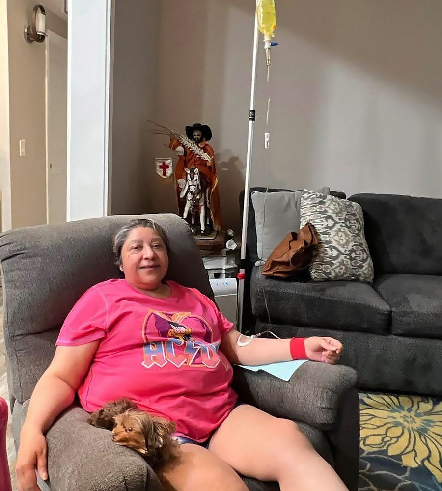 Woman in pink shirt receiving IV treatment, sitting in recliner, dog on lap.