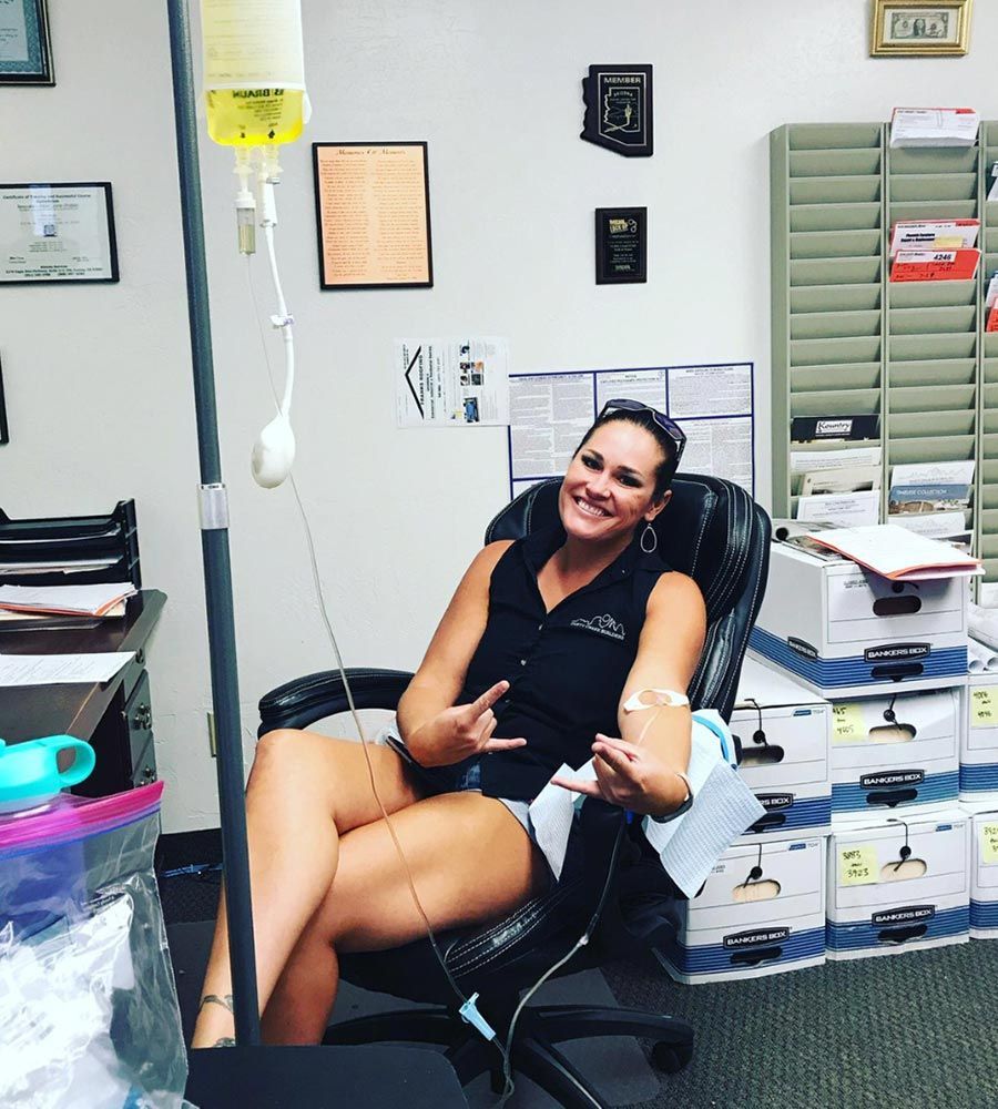 A person sits in an office chair making rock-on hand signs while receiving intravenous fluids from a hanging IV bag.