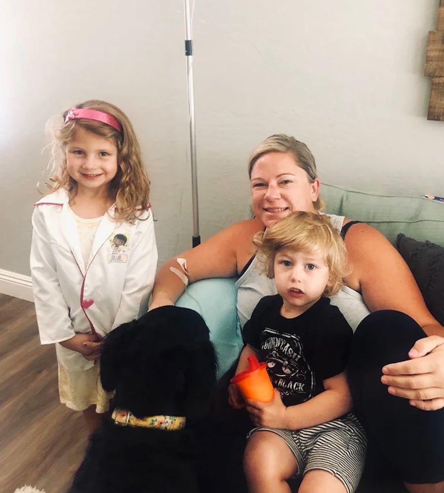 Smiling mother receiving IV treatment at home, sitting on the couch with her two young children and a large black dog.