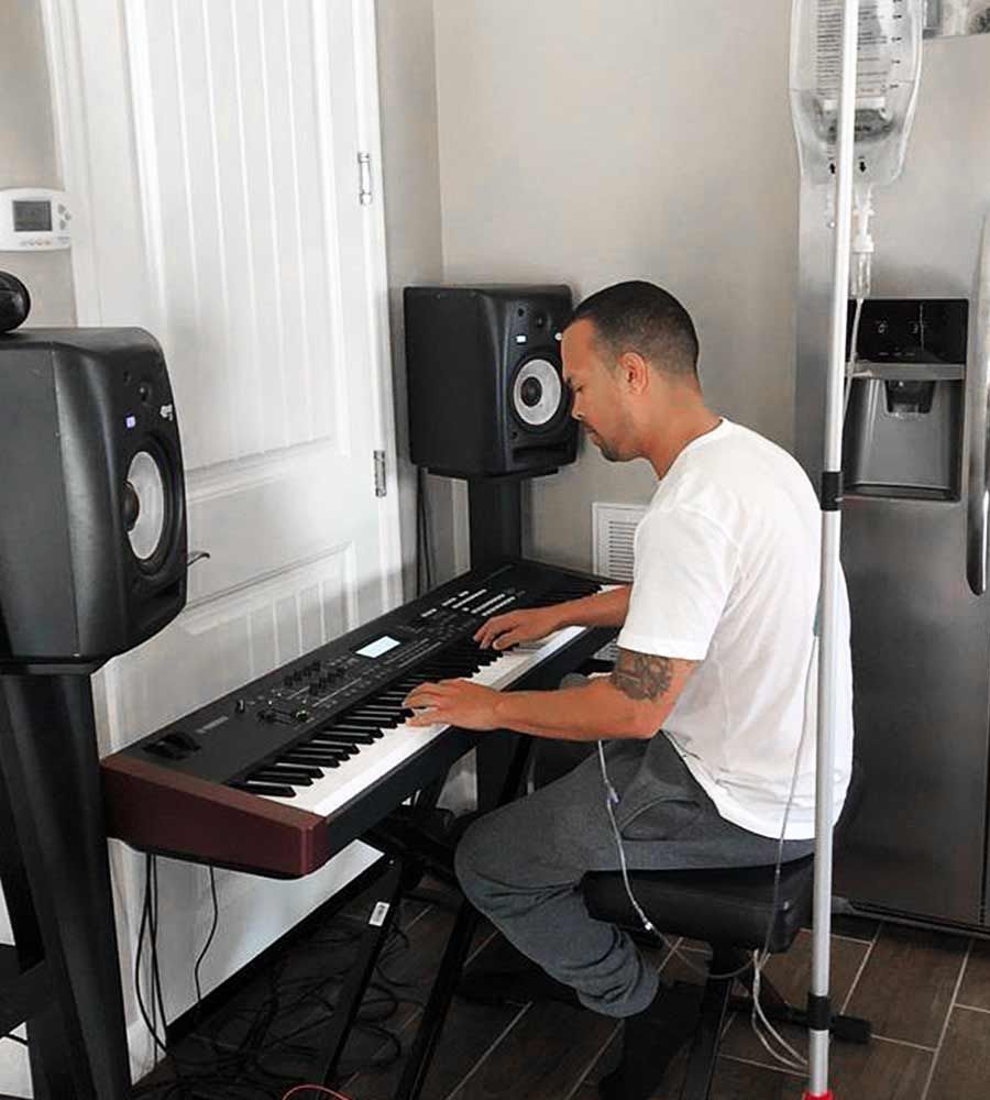 A musician sits at his keyboard/piano receiving mobile IV therapy for performance enhancement or wellness while working in his home studio.