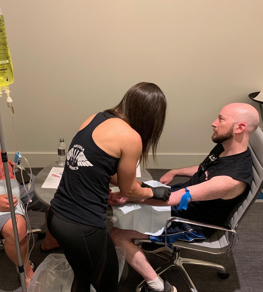Registered nurse administering a recovery or wellness IV drip infusion to a male patient sitting in an armchair, demonstrating convenient in-home IV services.