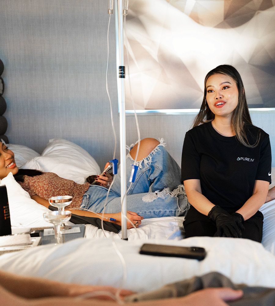Professional mobile IV therapist sitting and conversing with a female client who is relaxing and receiving an IV hydration drip on a hotel bed, emphasizing attentive and personalized service.