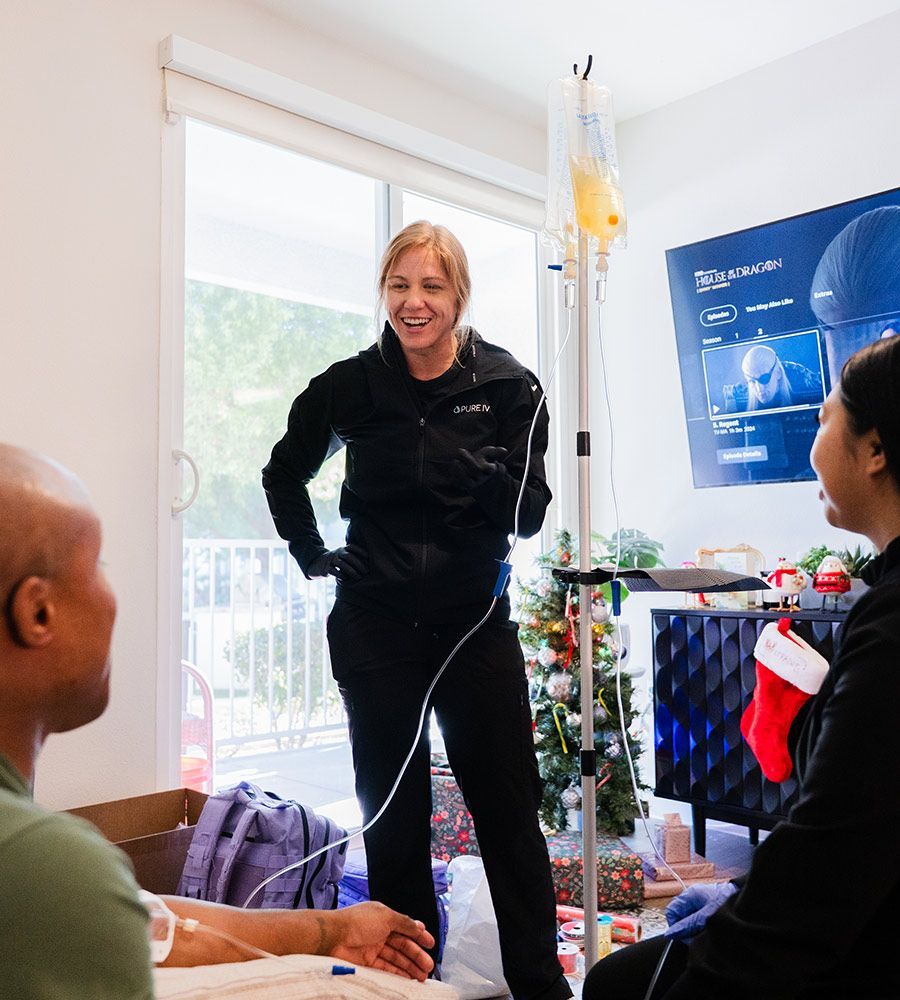 Smiling PURE IV nurse (Jaret) having a cheerful conversation with a couple in their living room, demonstrating friendly and professional at-home mobile IV hydration service near a Christmas tree.