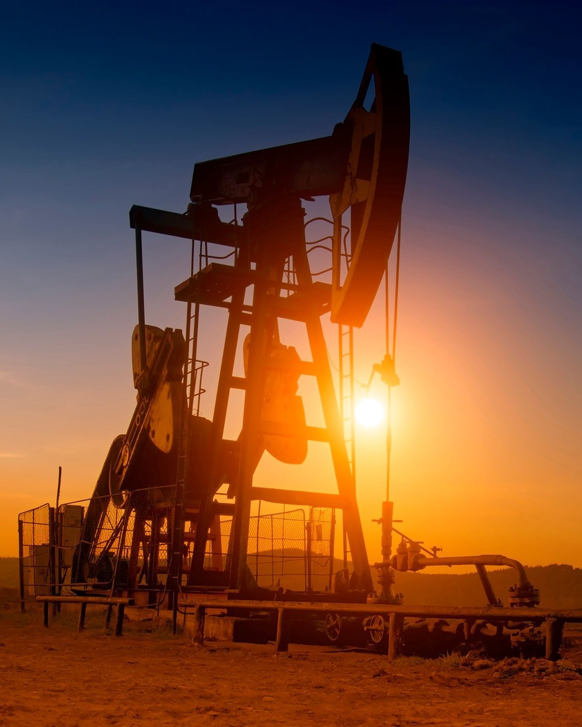 A pumpjack silhouette stands against a bright, orange sunset sky over a dusty landscape.