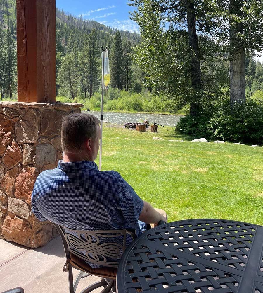 A man sitting on a patio overlooking a scenic river and forest while receiving an IV vitamin treatment.