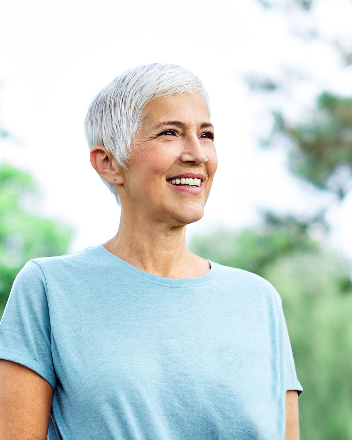A smiling person with short grey hair wearing a light blue t-shirt, looking away against a soft, green outdoor background.