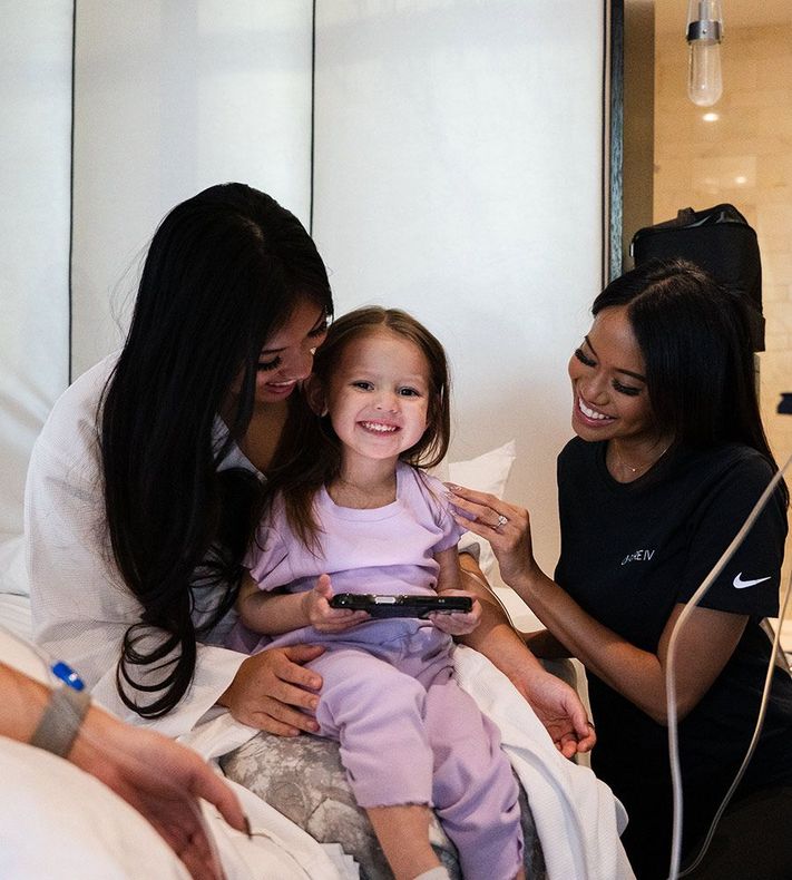 Two women and a child in a hotel room. The child smiles while holding a device. One woman touches the child.