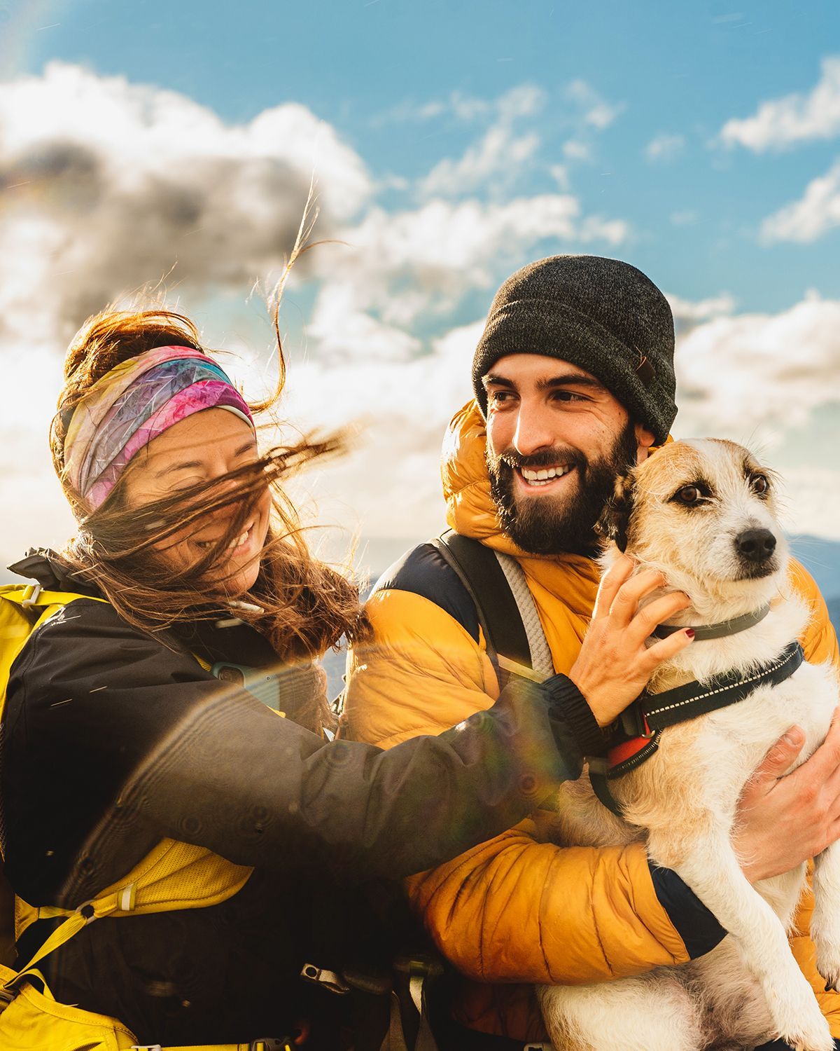 A couple in outdoor gear with windblown hair smiles while petting their dog on a mountain summit under a cloudy sky.