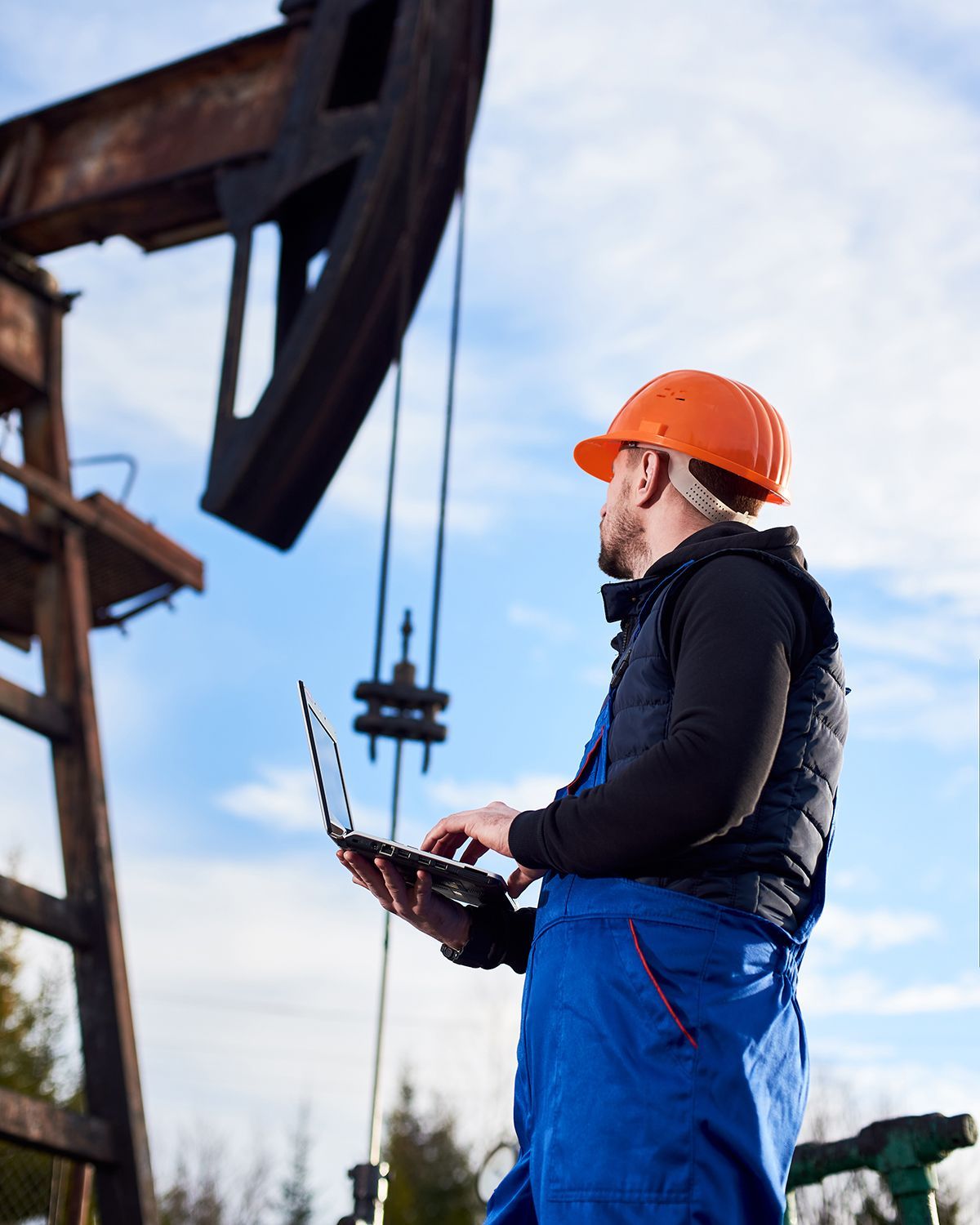 A worker in a hard hat and blue coveralls uses a laptop in front of an industrial oil pump jack against a cloudy sky.