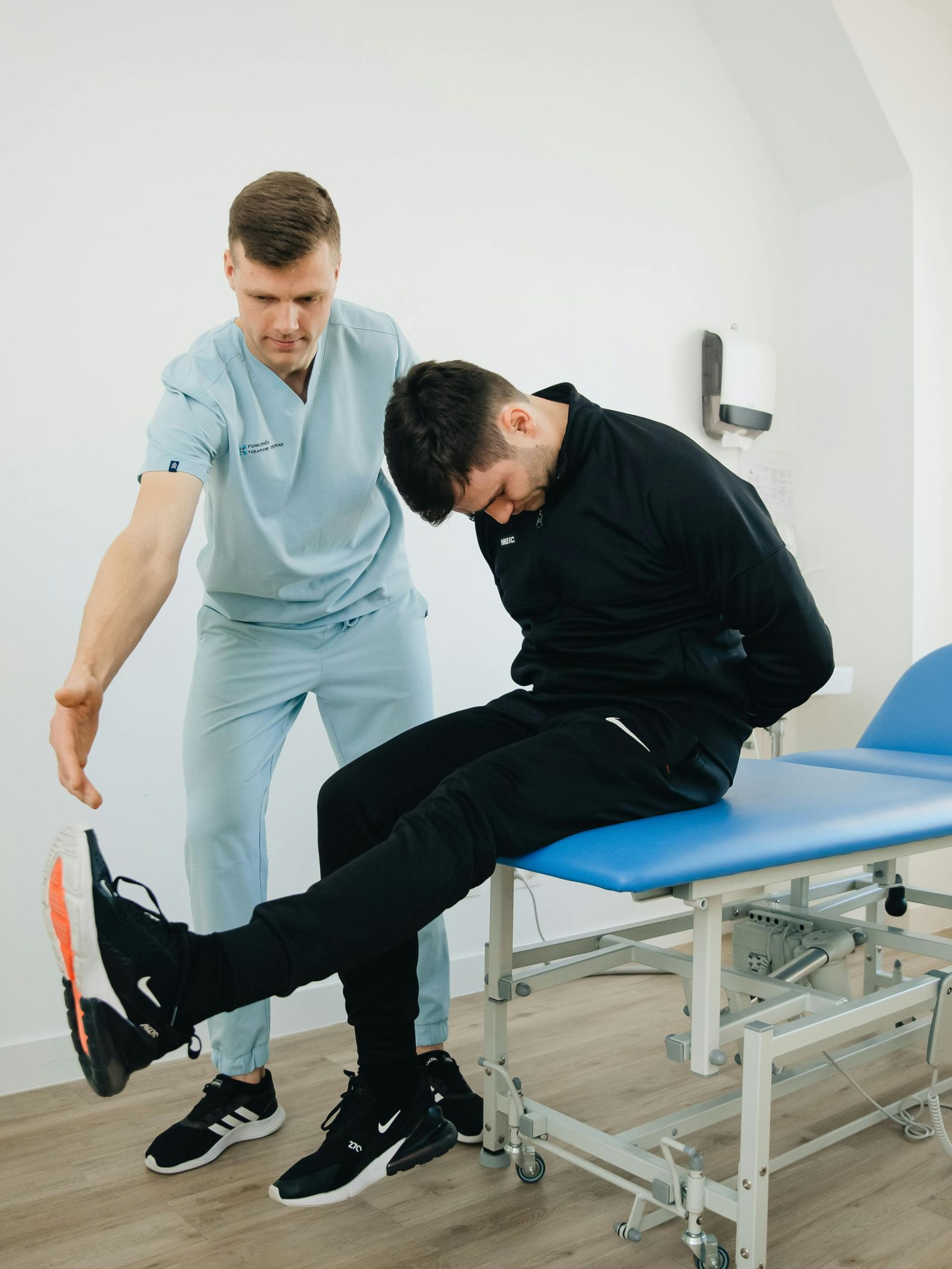A physical therapist guides a patient in a seated leg extension stretch on a clinic exam table.