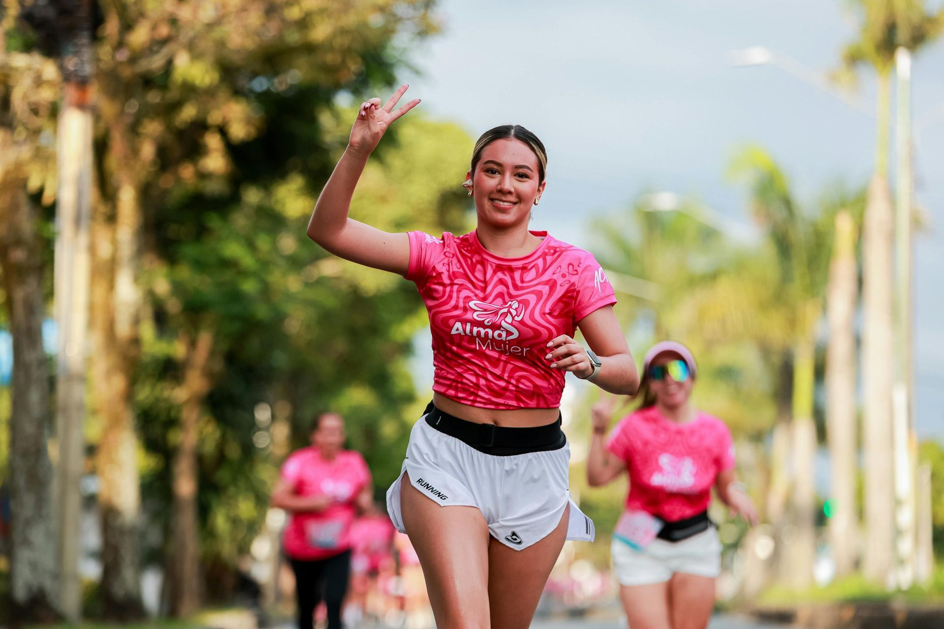 Woman in pink shirt and white shorts raises two fingers while running a race.