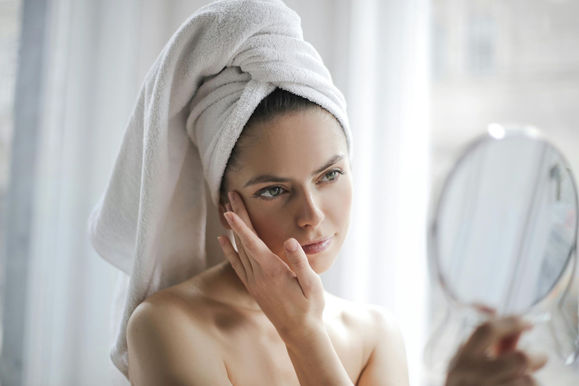 Woman with a towel on her head examines her face in a mirror. She's indoors near a window.