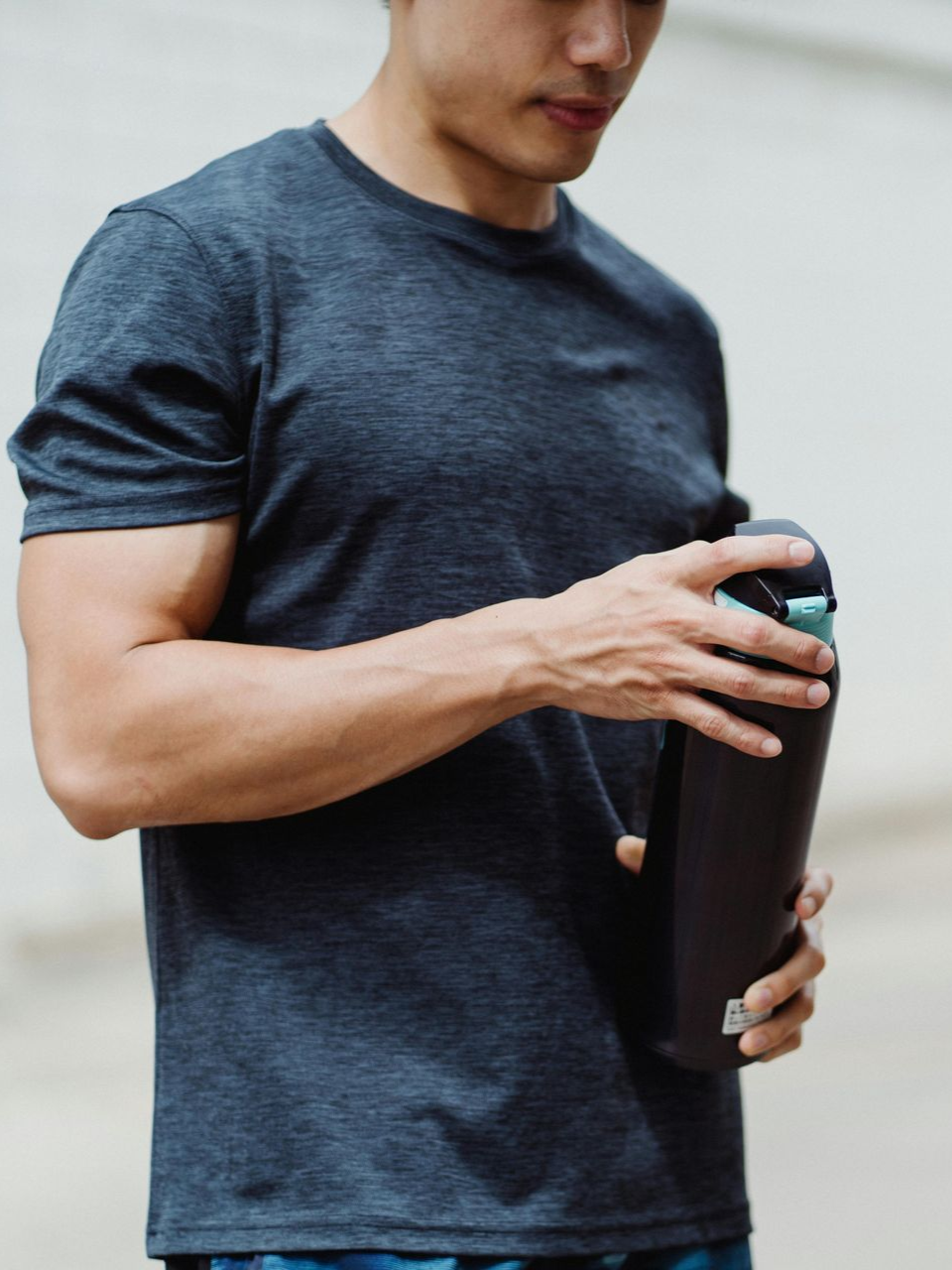 A person in a gray t-shirt stands outdoors, holding a black water bottle while opening its lid.