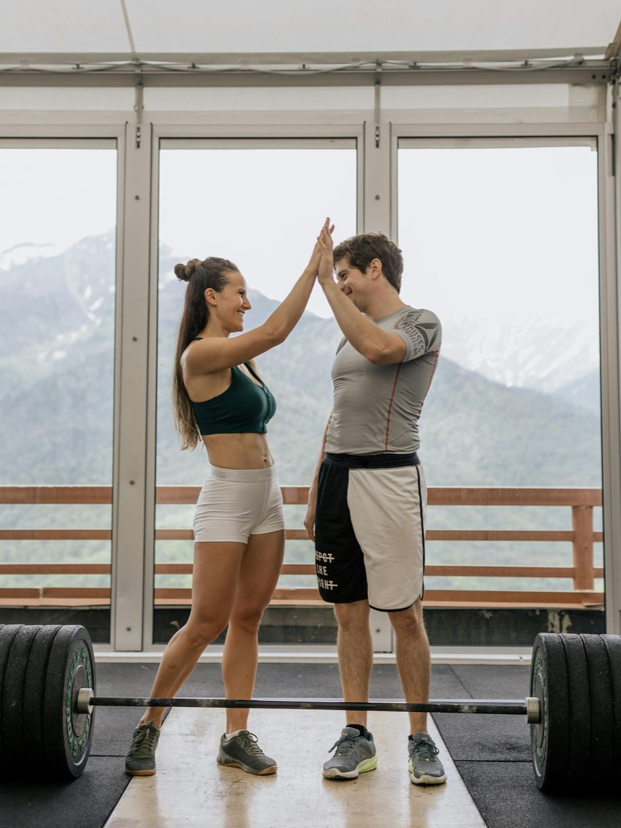 Two people in athletic wear high-five in a gym with a large window overlooking a mountain range.